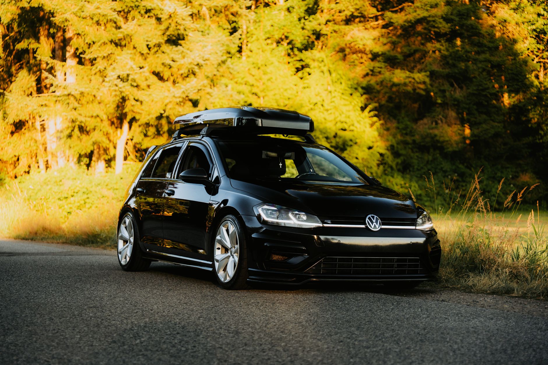 A modern black car with a roof box parked on a sunlit road surrounded by lush green forest.